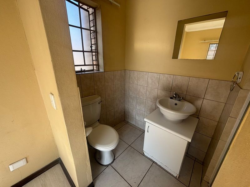 A compact bathroom featuring a standard toilet, vanity basin with storage, tiled flooring, and a window with security bars.