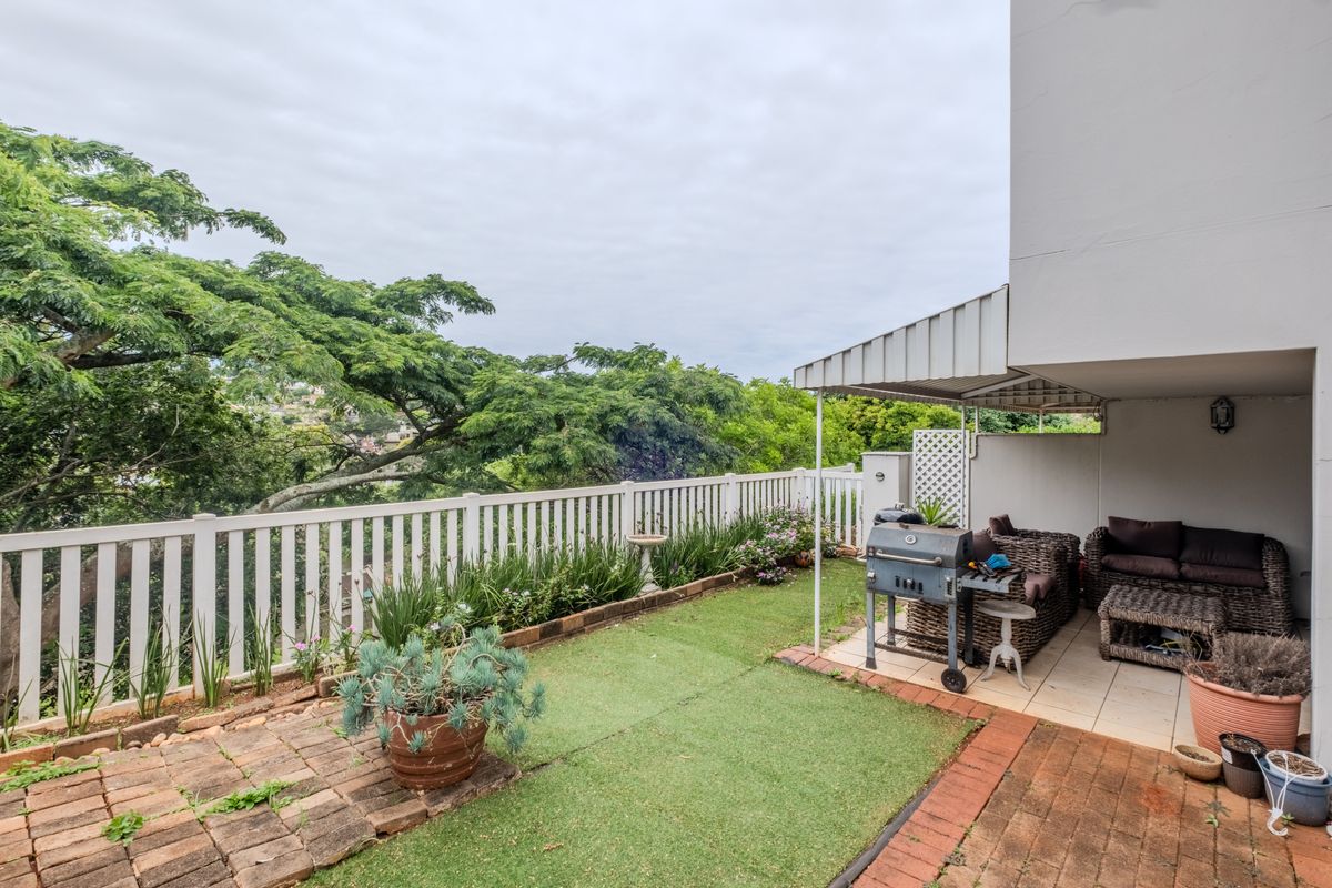 A covered patio area featuring tiled flooring, brick paving, and artificial grass, enclosed by a white picket fence with views of surrounding greenery.