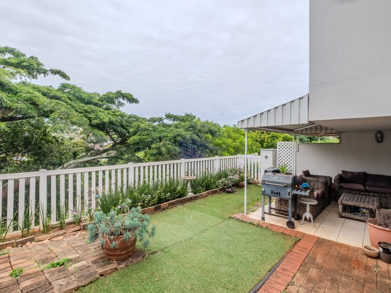 A covered patio area featuring tiled flooring, brick paving, and artificial grass, enclosed by a white picket fence with views of surrounding greenery.