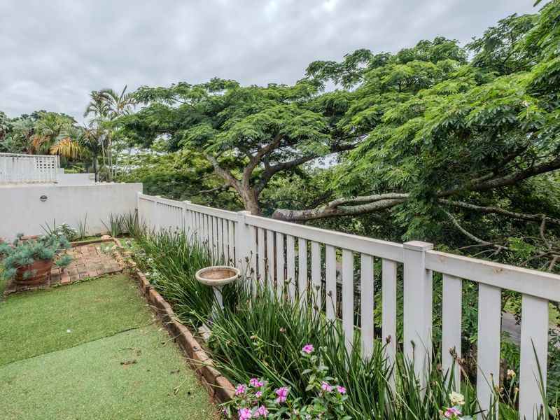 A private balcony area featuring a white picket fence, brick-edged garden beds, and views of mature trees, offering a peaceful outdoor space.