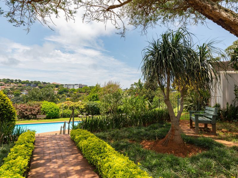 A well-maintained outdoor pool area featuring a brick-paved pathway, lush landscaped gardens, and scenic views of the surrounding neighborhood.