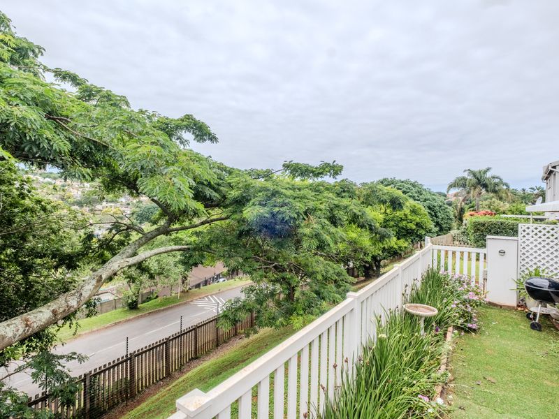 An elevated patio area featuring a white picket fence, overlooking a lush green landscape and the street below.
