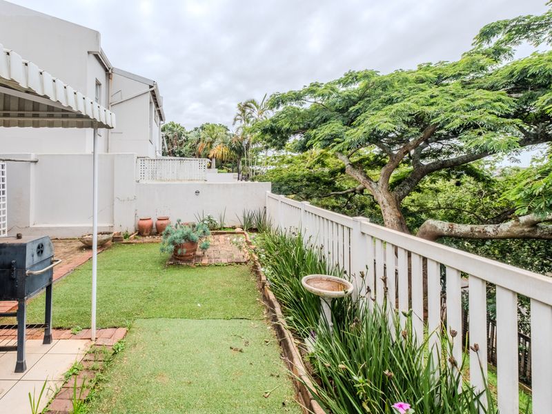 A private outdoor patio area featuring a retractable awning, paved sections, artificial turf, and a timber fence overlooking mature trees.