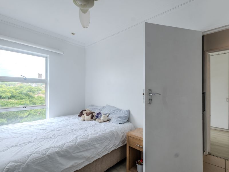 A bright bedroom featuring a ceiling fan, built-in cupboards, and a large window with views of the surrounding greenery.