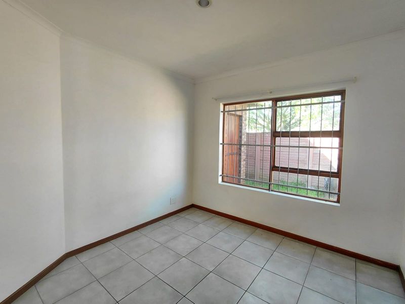 A bright, empty bedroom featuring neutral tiled flooring, clean white walls, and a window fitted with security bars.