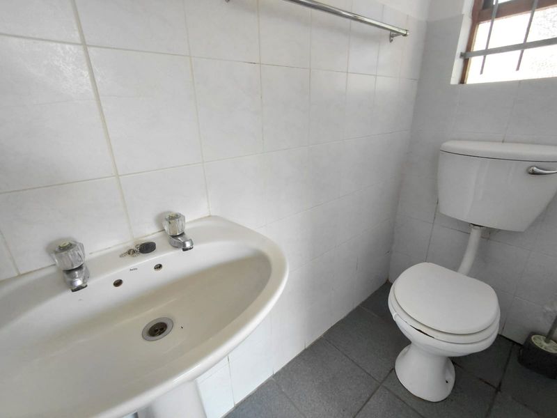 A functional bathroom featuring a pedestal basin, toilet, tiled walls and flooring, and a window with security bars.