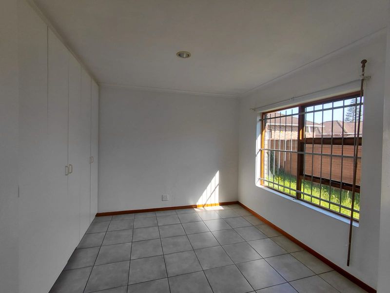 A bright, empty bedroom featuring neutral tiled flooring, built-in cupboards, and large windows fitted with security bars.