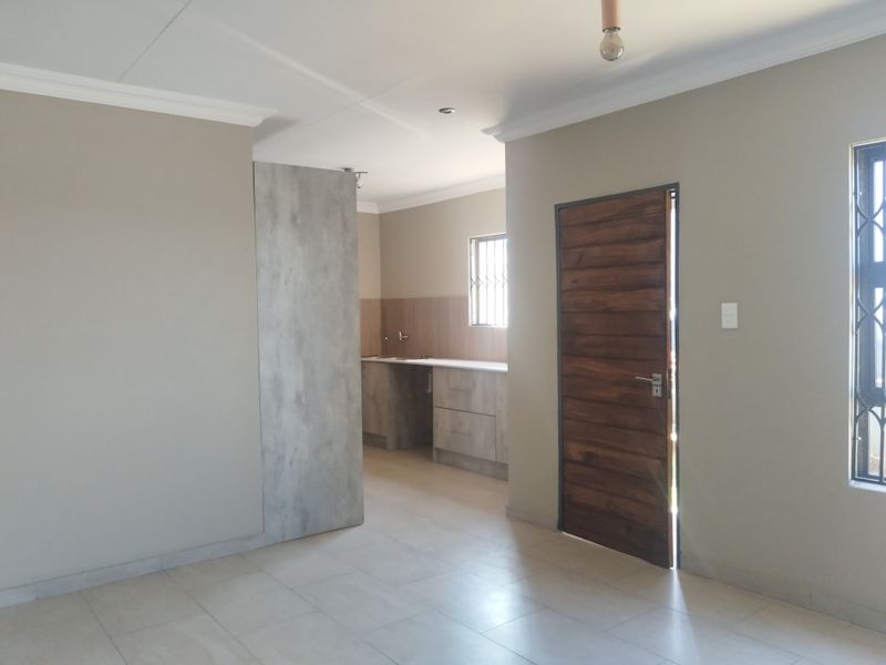 An open-plan living area featuring tiled floors, neutral walls, and a functional kitchen space with built-in cabinetry and sink.