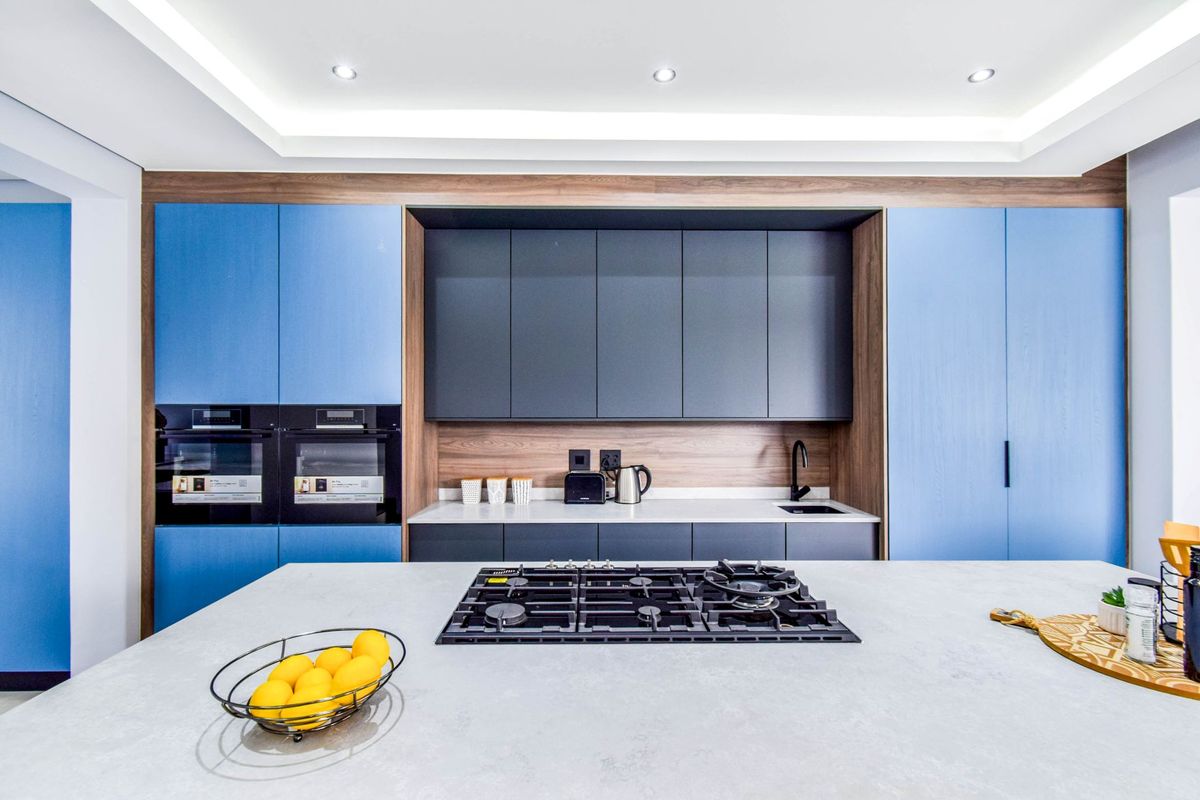 Modern kitchen featuring a central island with a built-in gas hob, sleek blue and charcoal cabinetry, integrated ovens, and high-quality stone countertops.