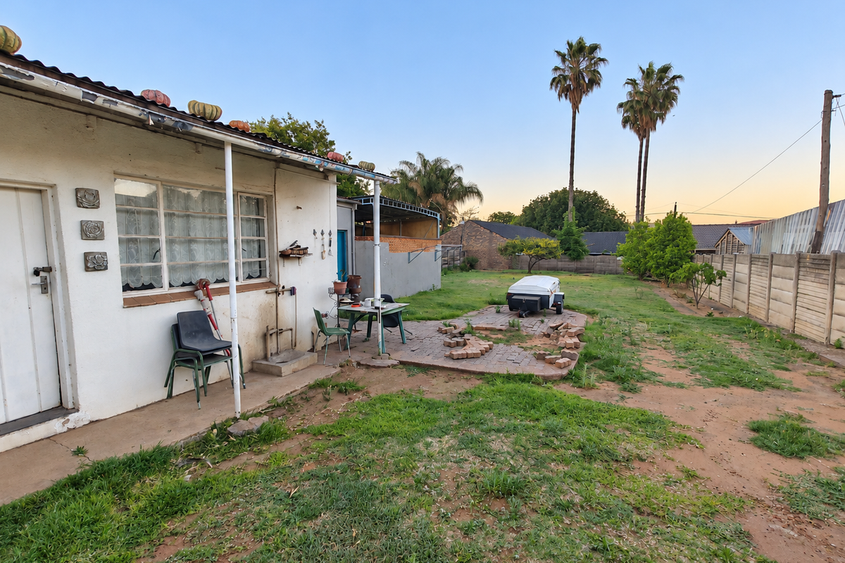 Rear exterior view of the property featuring a paved patio area, lawn, and perimeter walling, showing signs of wear and maintenance requirements.