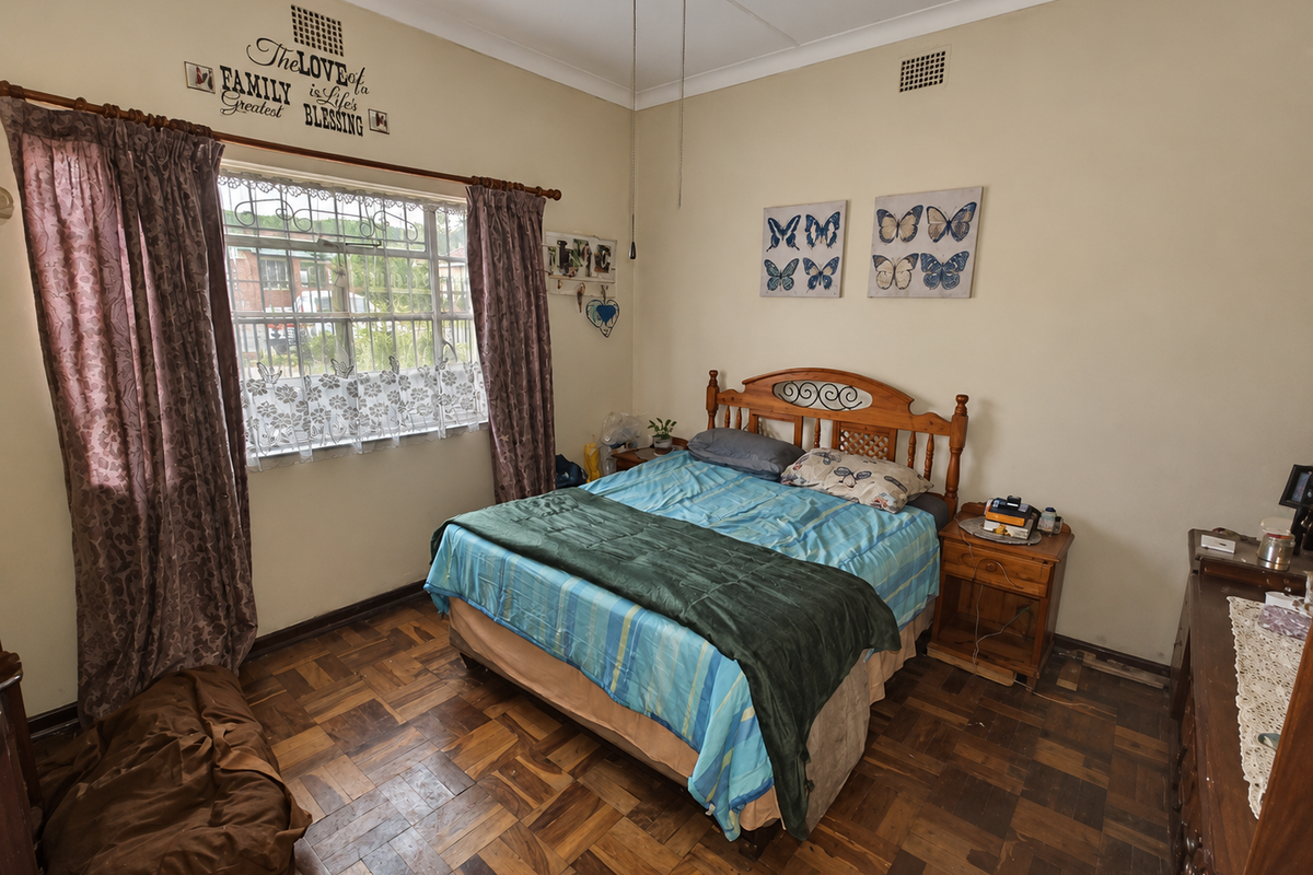 A bedroom featuring traditional parquet flooring, large window with security bars, and ceiling ventilation, offering a functional space with potential for cosmetic updates.