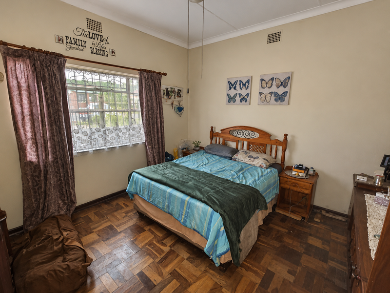 A bedroom featuring traditional parquet flooring, large window with security bars, and ceiling ventilation, offering a functional space with potential for cosmetic updates.
