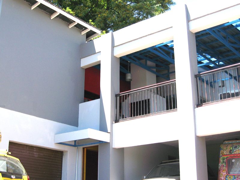 Modern two-story residential facade featuring a balcony with metal railings, exposed blue roof trusses, and a ground-level garage entrance.