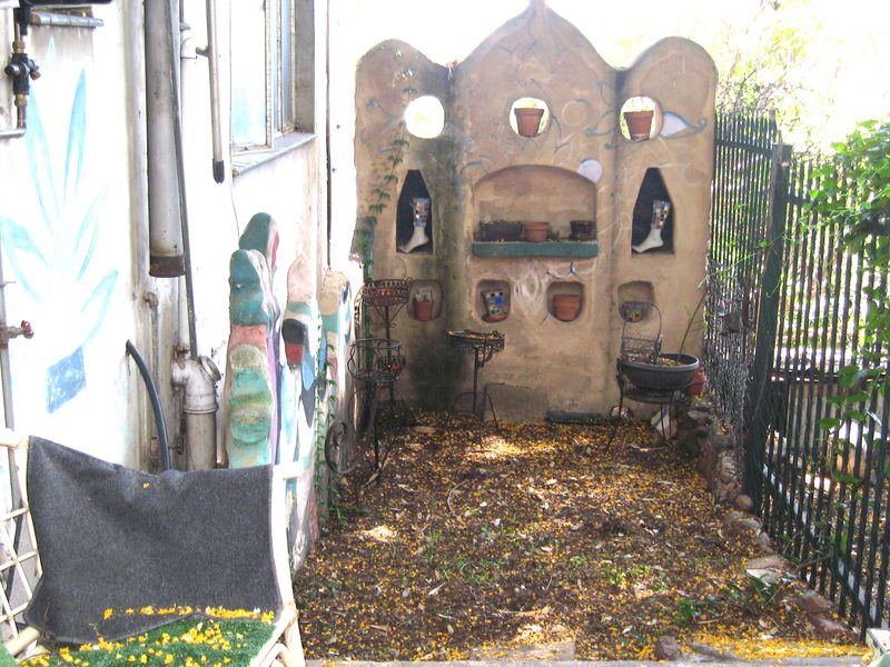 A small, enclosed garden area featuring a decorative masonry wall with built-in niches, metal fencing, and weathered exterior wall finishes.