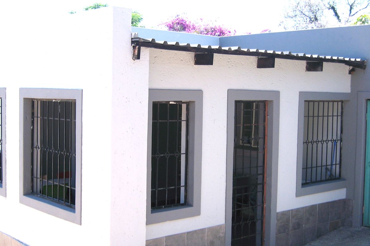 Exterior view of a building section featuring white stucco walls, grey trim, security bars on windows, and a corrugated metal roof.