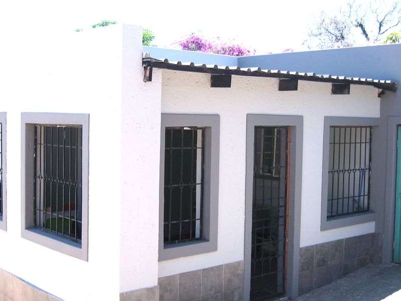 Exterior view of a building section featuring white stucco walls, grey trim, security bars on windows, and a corrugated metal roof.
