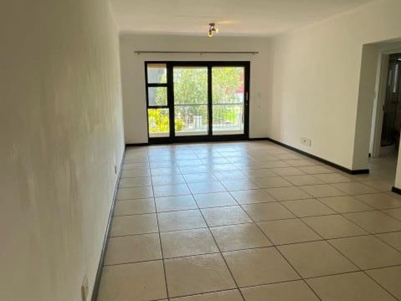 A spacious, empty living area featuring neutral tiled flooring, ceiling-mounted lighting, and a large sliding glass door providing access to the exterior.
