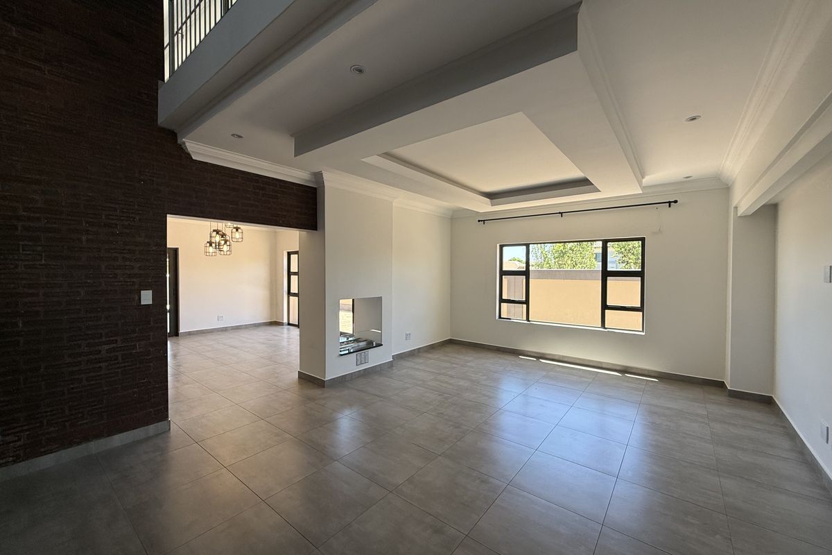Spacious open-plan living area featuring a double-volume ceiling, recessed lighting, tiled floors, and a striking exposed brick feature wall with a built-in fireplace.