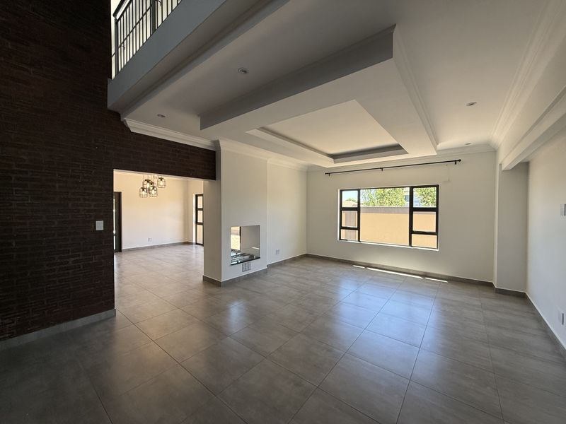 Spacious open-plan living area featuring a double-volume ceiling, recessed lighting, tiled floors, and a striking exposed brick feature wall with a built-in fireplace.