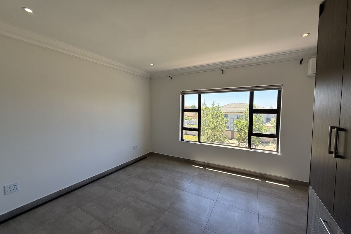 A bright, empty bedroom featuring modern tiled flooring, built-in cupboards, and large aluminium-framed windows providing natural light.