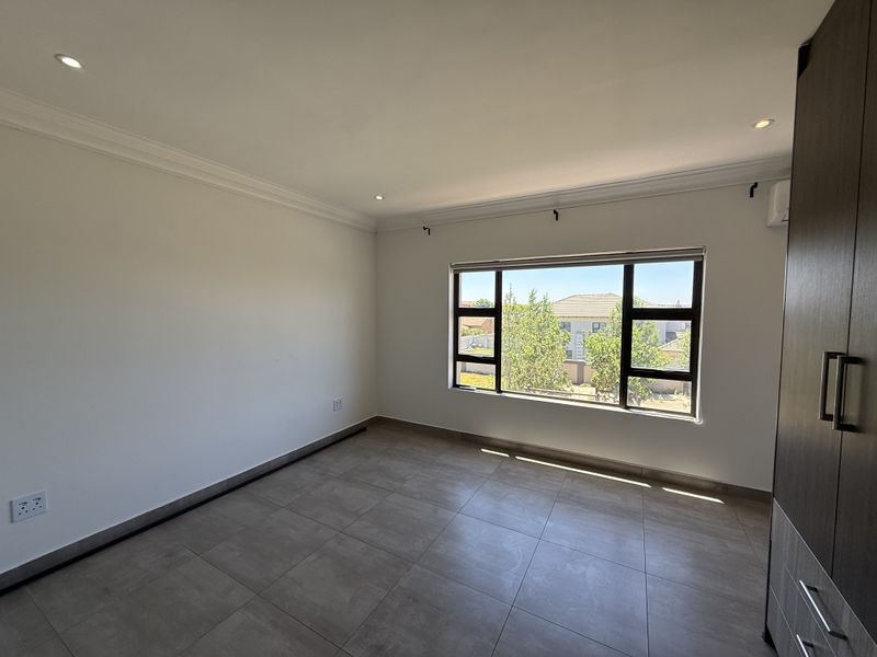 A bright, empty bedroom featuring modern tiled flooring, built-in cupboards, and large aluminium-framed windows providing natural light.