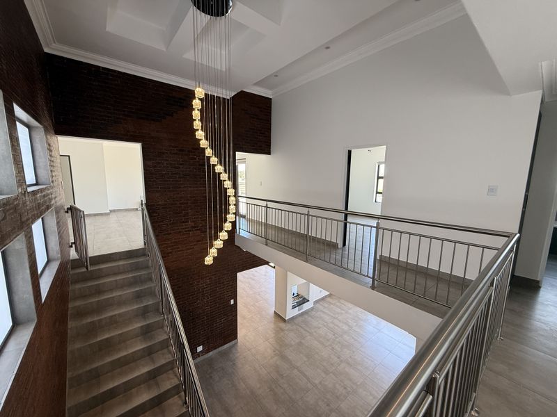 Spacious double-volume landing area featuring a modern staircase, stainless steel balustrades, an exposed brick feature wall, and an elegant pendant chandelier.