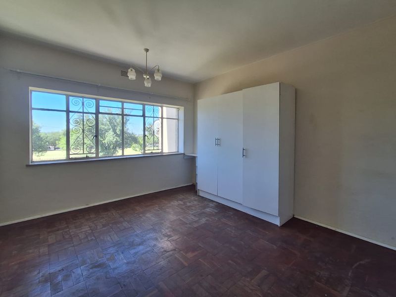A spacious bedroom featuring original parquet flooring, a built-in wardrobe, and large windows with security bars, offering a blank canvas for renovation.