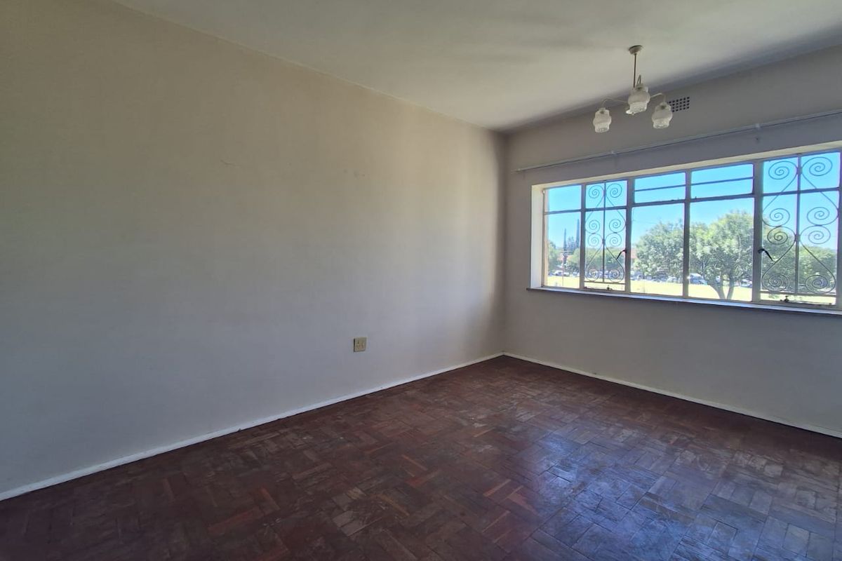 A spacious, empty bedroom featuring original parquet flooring, a ceiling light fixture, and large windows with security bars.