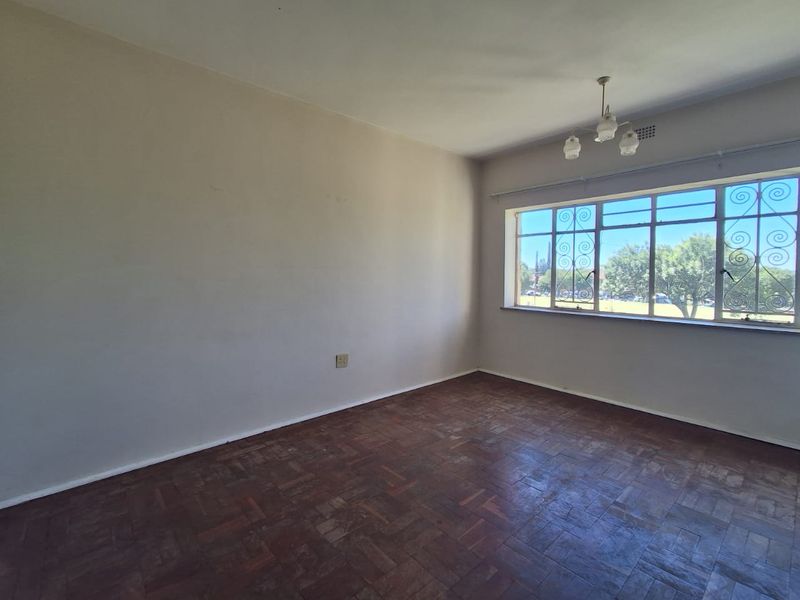 A spacious, empty bedroom featuring original parquet flooring, a ceiling light fixture, and large windows with security bars.
