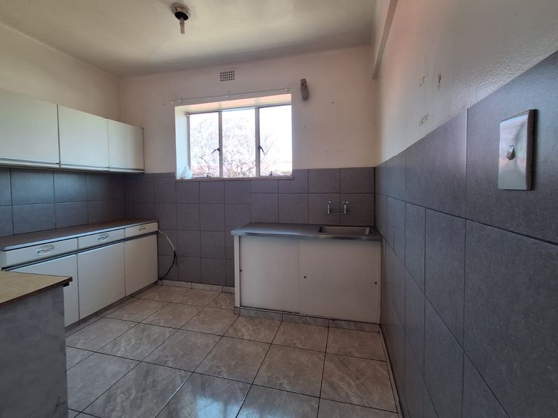 A functional, empty kitchen featuring tiled flooring, grey tiled splashbacks, built-in cabinetry, and a stainless steel sink, ready for a modern update.