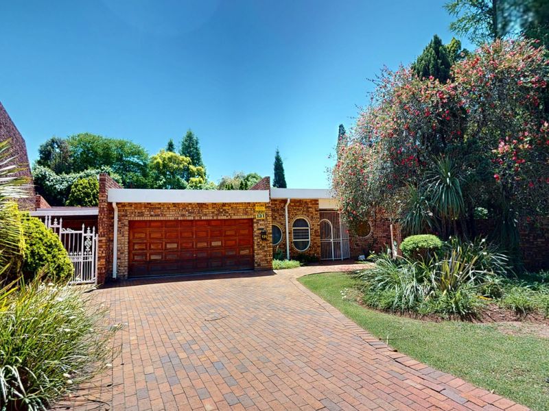 Front elevation of a brick house featuring a double garage, paved driveway, and established garden with mature trees.