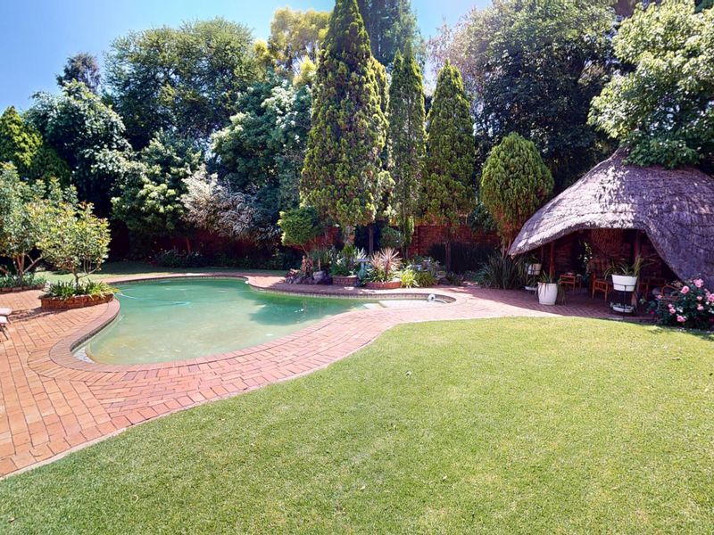 A spacious outdoor pool area featuring a swimming pool, brick paving, and a traditional thatched lapa set within a mature, landscaped garden.