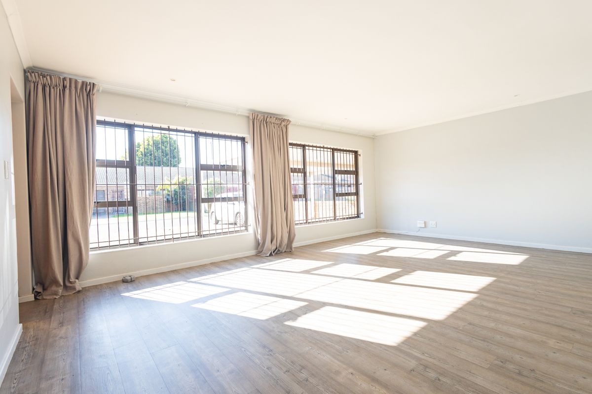 A bright and spacious living area featuring light-toned laminate flooring, large windows with security bars, and neutral wall finishes.