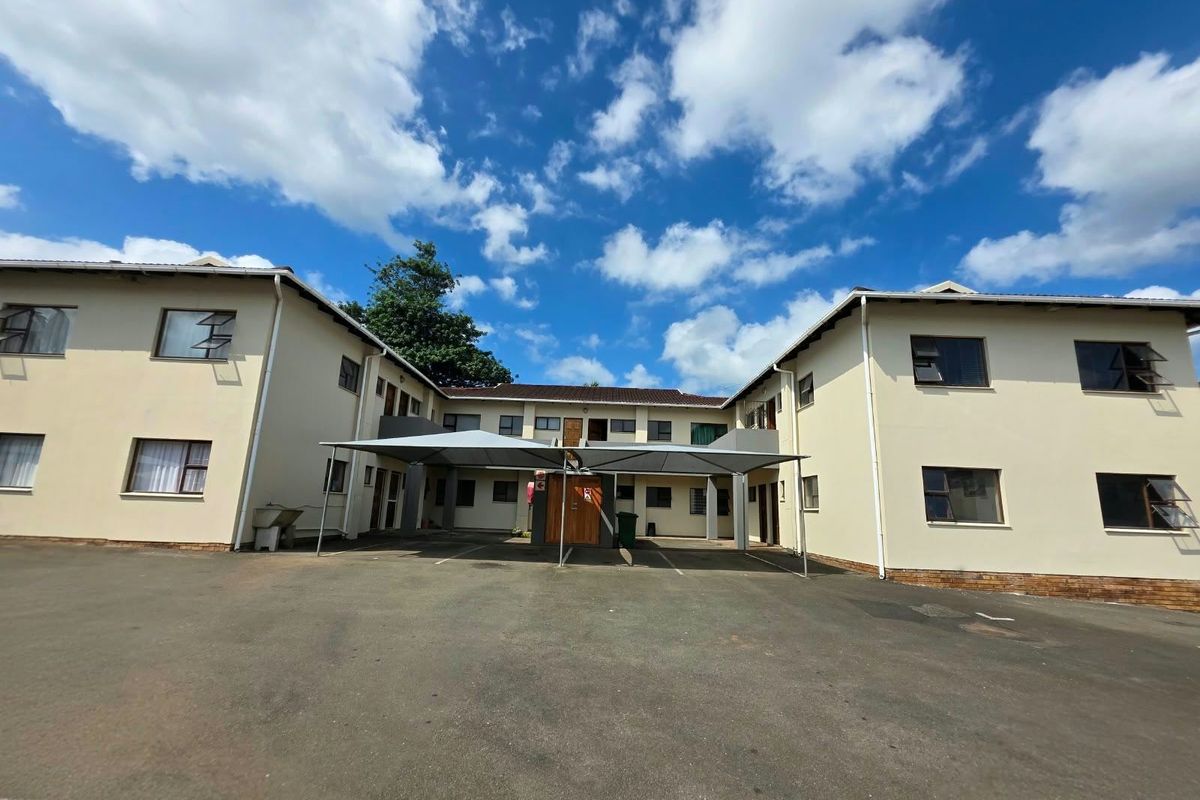 Front view of an apartment complex featuring a paved parking area with a shade cloth carport structure and multi-story residential units.