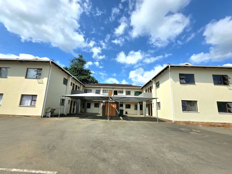 Front exterior view of the apartment complex featuring a paved driveway and covered parking bays under shade cloth structures.
