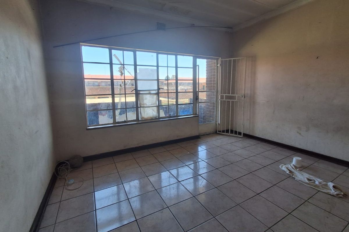 An empty living room featuring tiled flooring, a large steel-framed window, and a security gate, requiring cosmetic updates and wall repairs.