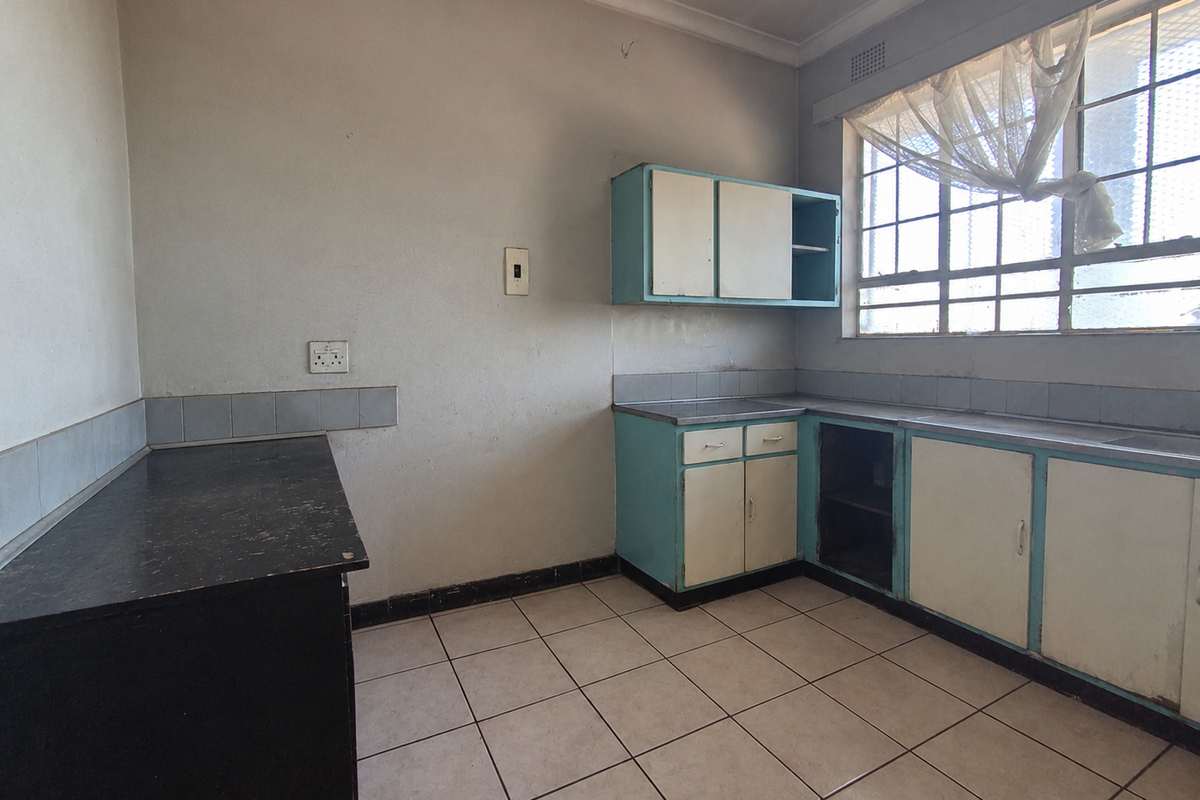 An empty, dated kitchen featuring original cabinetry, tiled flooring, and a window with security bars, offering significant potential for a modern renovation.