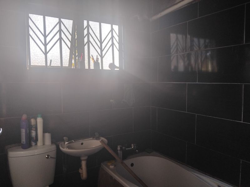 A bathroom featuring dark wall tiles, a built-in bathtub, a wall-mounted basin, and a toilet, with natural light entering through a window with security bars.