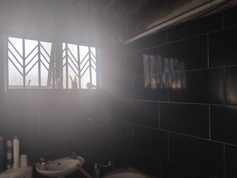 A bathroom featuring dark wall tiles, a pedestal basin, and a window with security bars, currently showing exposed ceiling and plumbing elements.