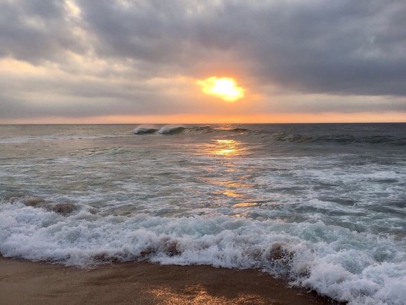 A scenic view of the ocean waves crashing on the sandy beach during a cloudy sunset.