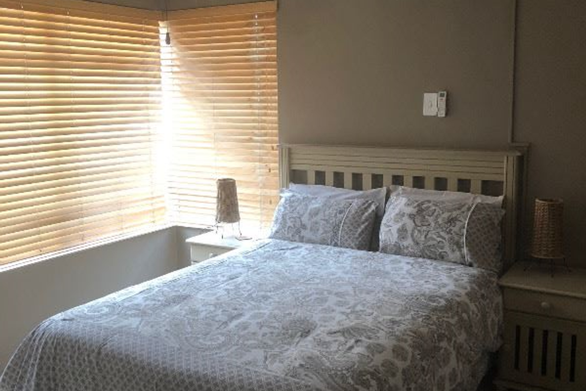 A functional bedroom featuring tiled flooring, a ceiling fan, and a wall-mounted split-system air conditioner, complemented by natural light through venetian blinds.