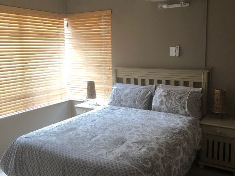 A functional bedroom featuring tiled flooring, a ceiling fan, and a wall-mounted split-system air conditioner, complemented by natural light through venetian blinds.