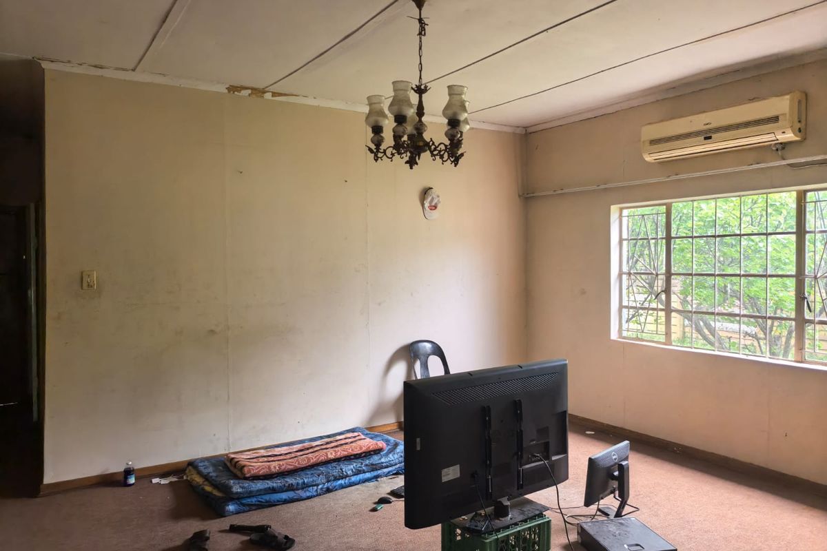 A spacious living room featuring a split-system air conditioner, security bars on the window, and a central chandelier, requiring cosmetic updates and flooring replacement.