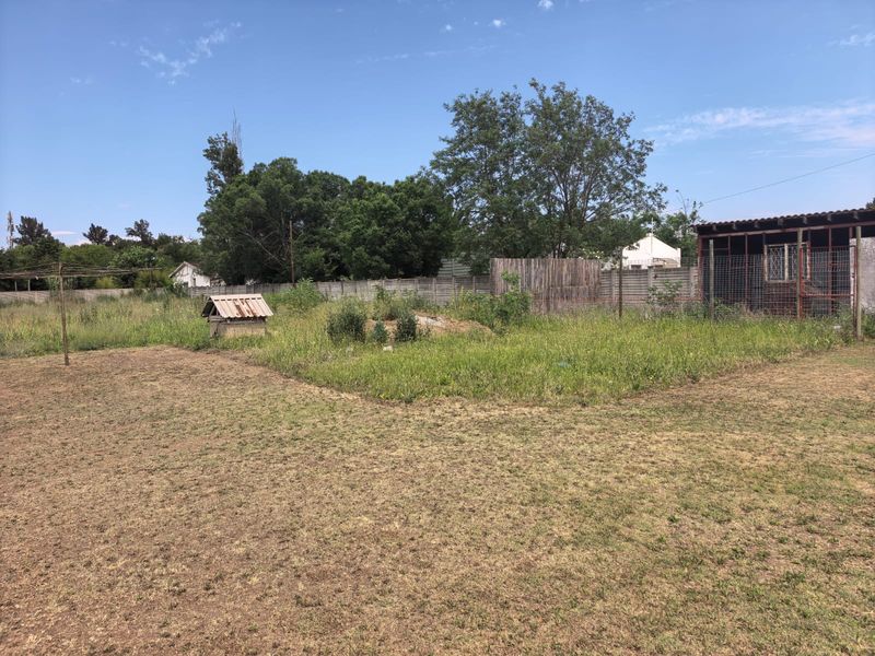 A large, unmaintained garden area featuring a precast concrete boundary wall, a small wellhead, and a covered outdoor structure in need of landscaping.