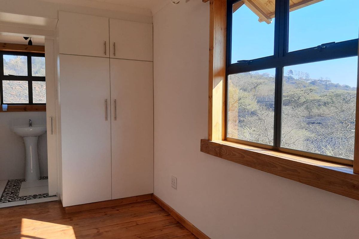A bright bedroom featuring timber flooring, built-in cupboards, and an en-suite bathroom with a pedestal basin, offering scenic views through timber-framed windows.