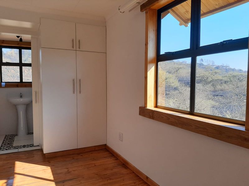 A bright bedroom featuring timber flooring, built-in cupboards, and an en-suite bathroom with a pedestal basin, offering scenic views through timber-framed windows.