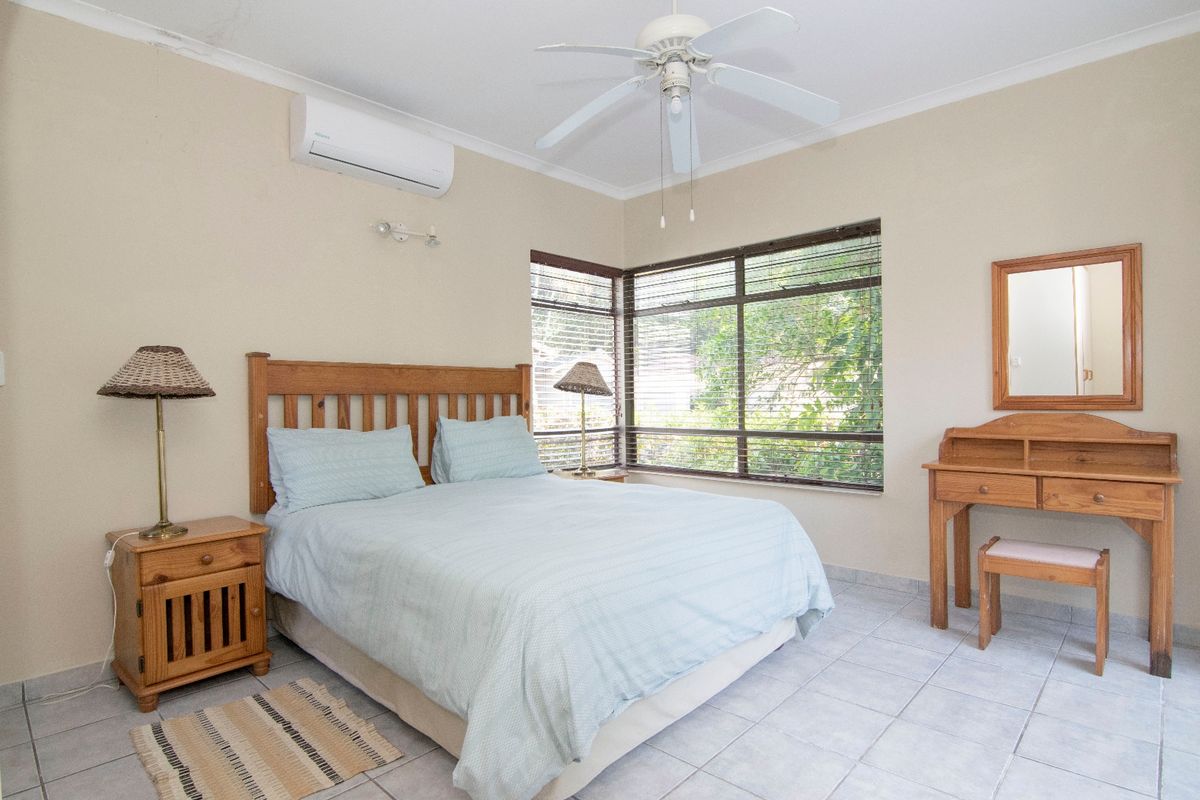 A well-lit bedroom featuring tiled flooring, a ceiling fan, and a split-system air conditioner, with large corner windows providing natural light.