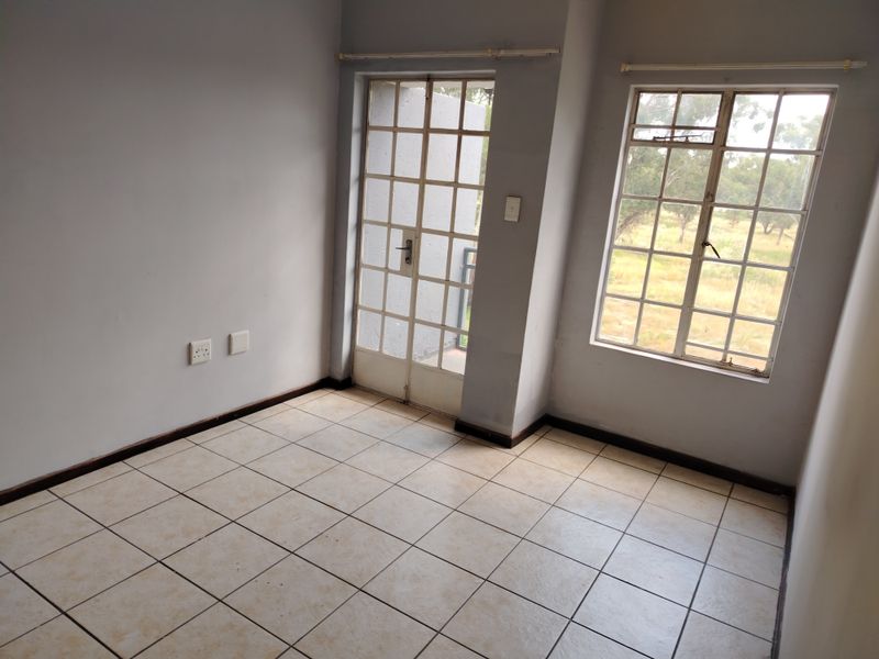 An empty living area featuring tiled flooring, a security gate leading outdoors, and windows with burglar bars, offering a blank canvas for renovation.