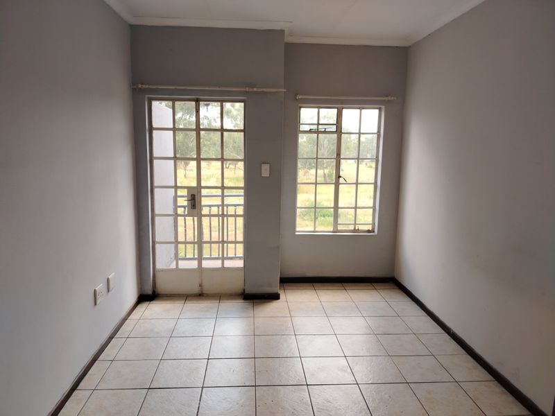 An empty living area featuring tiled flooring, a steel-framed glass door leading to a balcony, and a window with natural light.