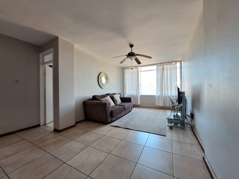 A bright living room featuring neutral tiled flooring, a ceiling fan, and large windows that provide ample natural light.